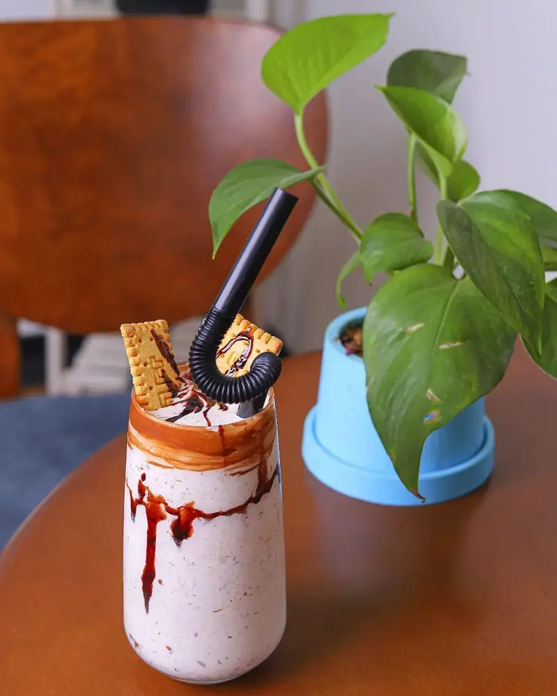 نوشیدنی خنک و خوشمزه کافه بینج با میلک شیک غلیظ، سس شکلات و بیسکوییت روی میز چوبی در محیط دنج کافه.