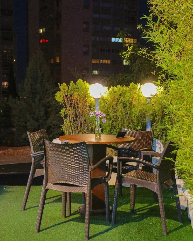 فضای باز دلنشین کافه بینج در شب، با میز چوبی و صندلی‌های حصیری، نورپردازی گرم، محیطی آرام برای لذت بردن از قهوه و دورهمی.