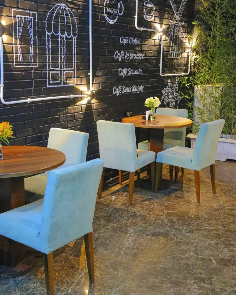 فضای کافه بینج با دیوار آجری مشکی، نقاشی‌های گچی، میزهای چوبی دنج و صندلی‌های پارچه‌ای روشن. محیطی آرام و دلنشین برای مشتریان.