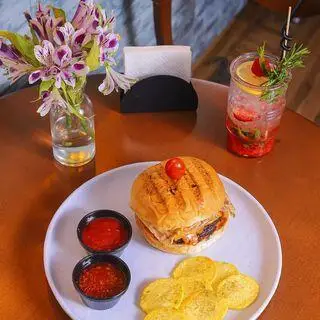 نمای نزدیک از غذای خوش طعم شامل همبرگر، چیپس و نوشیدنی میوه‌ای خنک در کافه بینج با چیدمان زیبا و گل‌های بنفش.