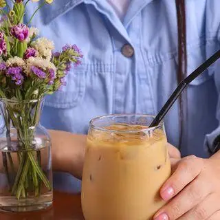 مشتری در کافه بینج، از نوشیدن قهوه سرد لاته با نی مشکی خود کنار گلدان گل‌ها در فضای آرام کافه لذت می‌برد.