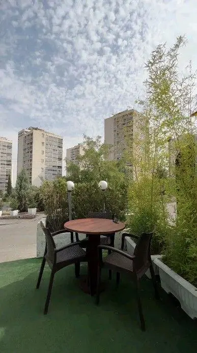 فضای باز کافه بینج با میز و صندلی چوبی، گیاهان سبز و بامبو، محیطی دلنشین در دل شهر با آسمان ابری و ساختمان‌های بلند.