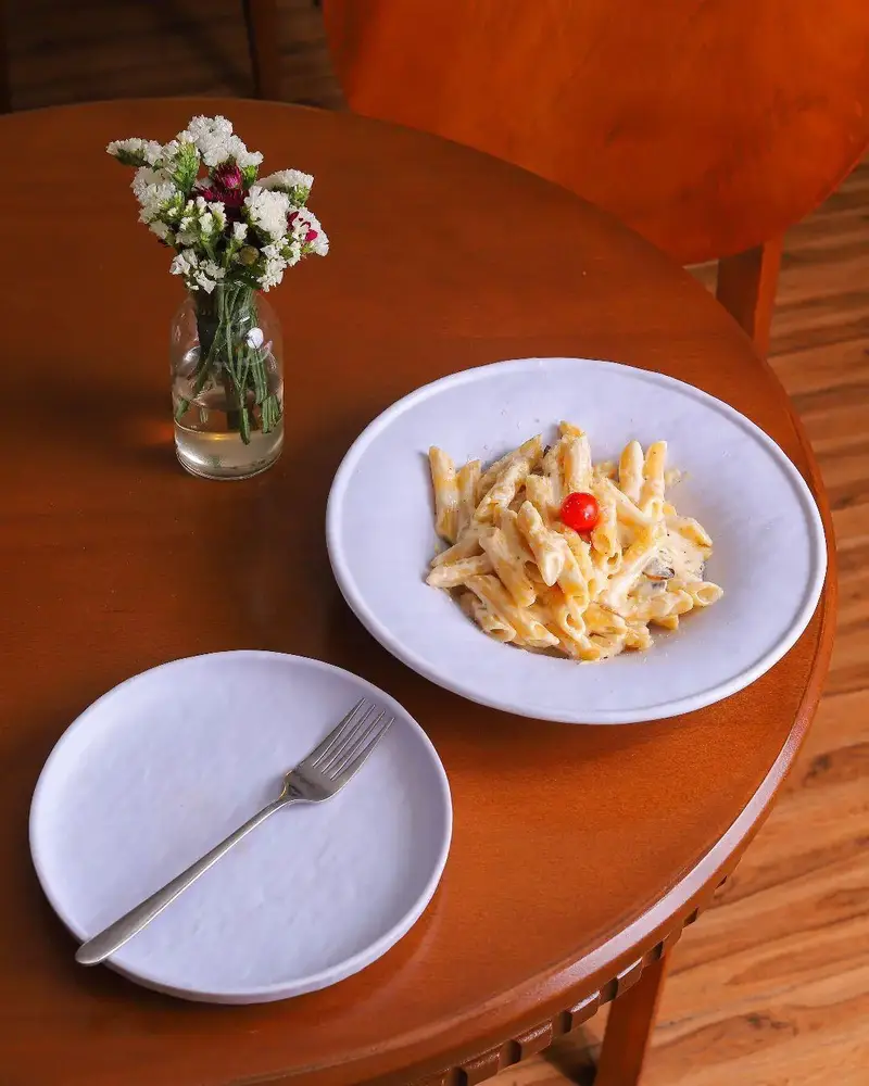 پاستا پنه خامه‌ای اشتهاآور در کافه بینج. میز چوبی با گلدان گل و بشقاب و چنگال. تجربه‌ای دلپذیر از غذای با کیفیت در محیط دنج.