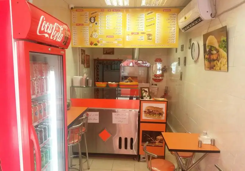 تصویری از داخل فست فود احسان با منوهای بزرگ، یخچال کوکاکولا، پیشخوان غذا و میز و صندلی‌های نارنجی و دکوراسیون گرم و دلنشین.