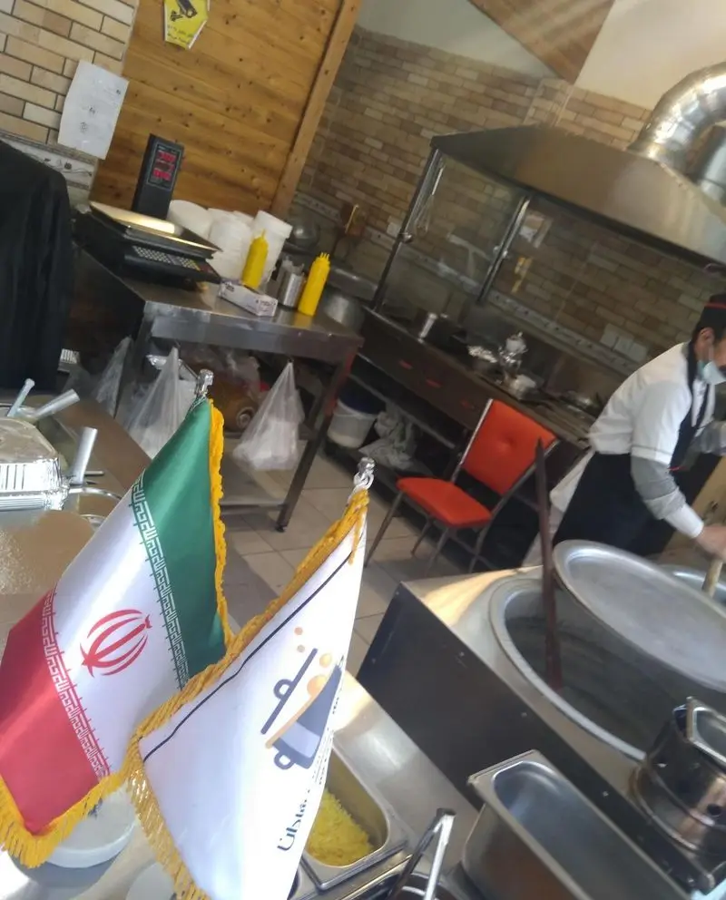 نمای داخلی رستوران غذای خونگی فود شادان با پرچم ایران، آشپز مشغول پخت و پز در آشپزخانه صنعتی و تمیز.