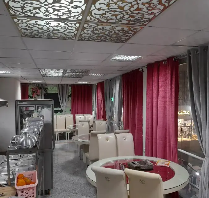 نمای داخلی رستوران فود شادان با میزهای گرد، صندلی‌های سفید، بوفه غذاخوری و سقف دکوراتیو، محیطی دلپذیر برای غذای خانگی.