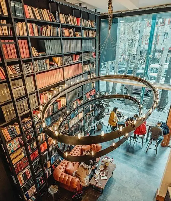 کتابخانه و کافه لمیز باغ فردوس با قفسه‌های بلند کتاب، لوستر دایره‌ای بزرگ و فضایی دلنشین برای مطالعه و قهوه.