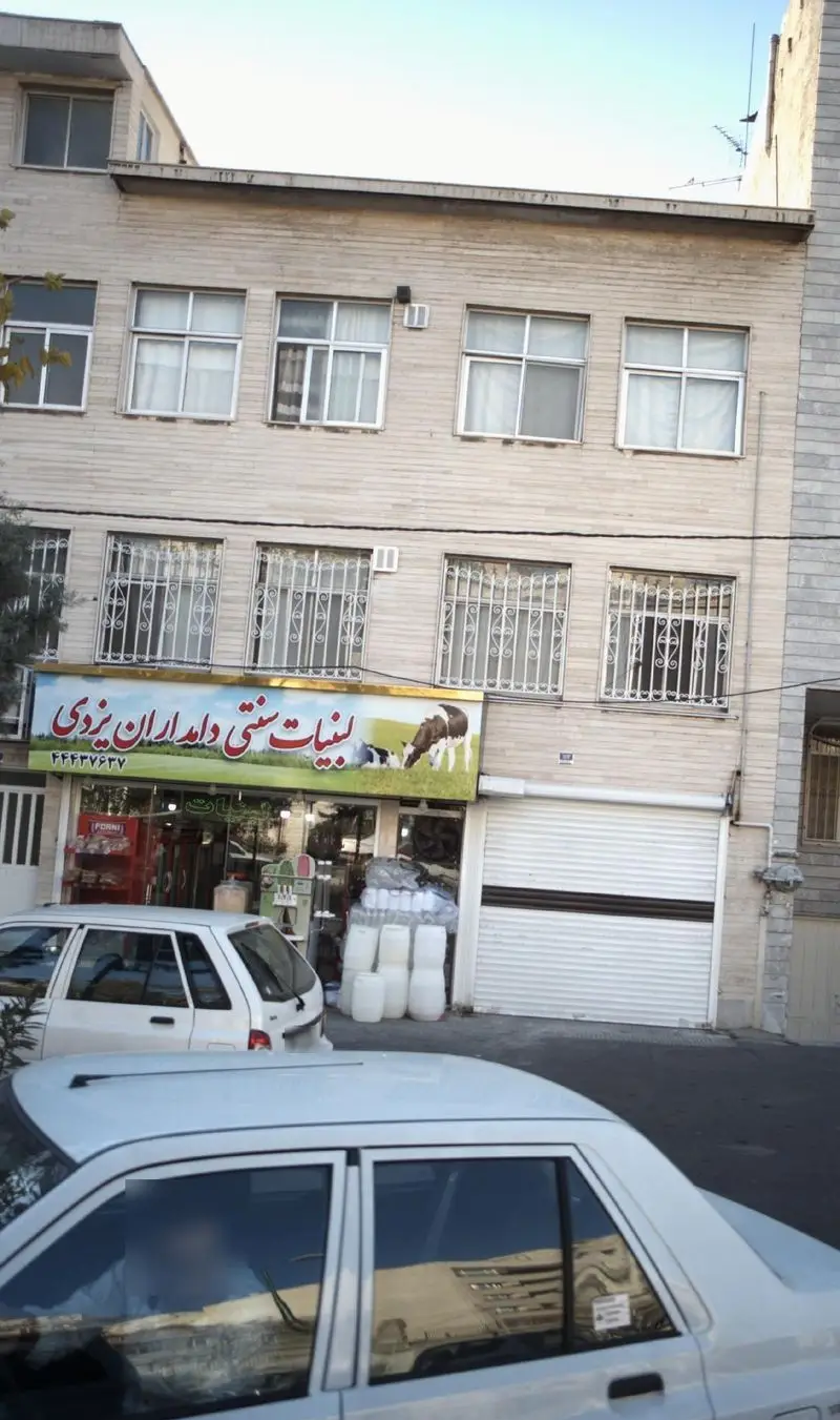 نمای بیرونی فروشگاه لبنیات سنتی دامداران یزدی با تابلوی بزرگ و تصویر گاوها، همراه با خودروها و دبه‌های محصولات لبنی.