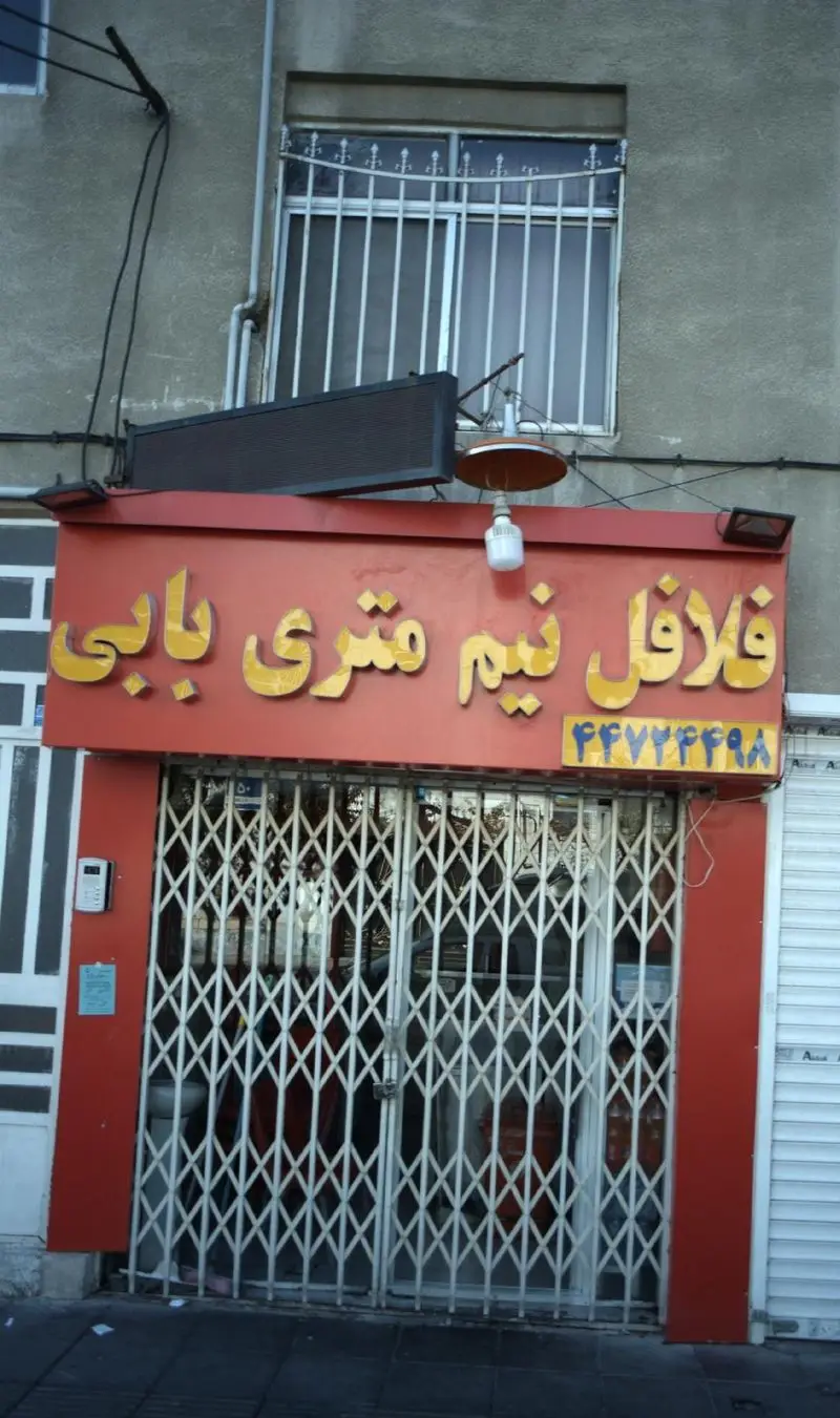 نمای بیرونی مغازه فلافل فروشی نیم متری بابی با تابلو بزرگ قرمز رنگ، نوشته‌های زرد فارسی و شماره تلفن ۴۴۷۷۲۴۹۸، درهای بسته فلزی.