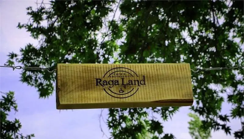 تابلو چوبی شیک رستوران فرنگی و کافه راگا لند (Raga Land) معلق در فضای دلنشین و سرسبز، احاطه شده با شاخ و برگ درختان.