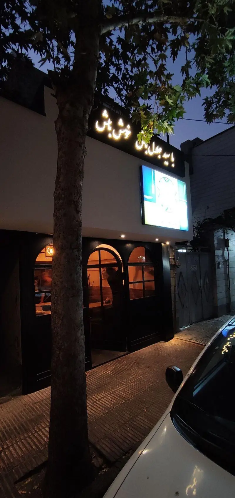 رستوران سید جلال در شب، با تابلوی نورانی "سید جلال خوش باش"، پنجره‌های قوسی روشن و گرم، و درختی که جلوی آن قرار دارد.