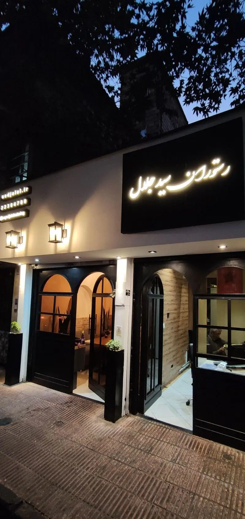 نمای شبانه رستوران سید جلال در تهران با تابلوی نورانی، درب‌های ورودی قوسی شیک و نورپردازی گرم، آماده پذیرایی از میهمانان.