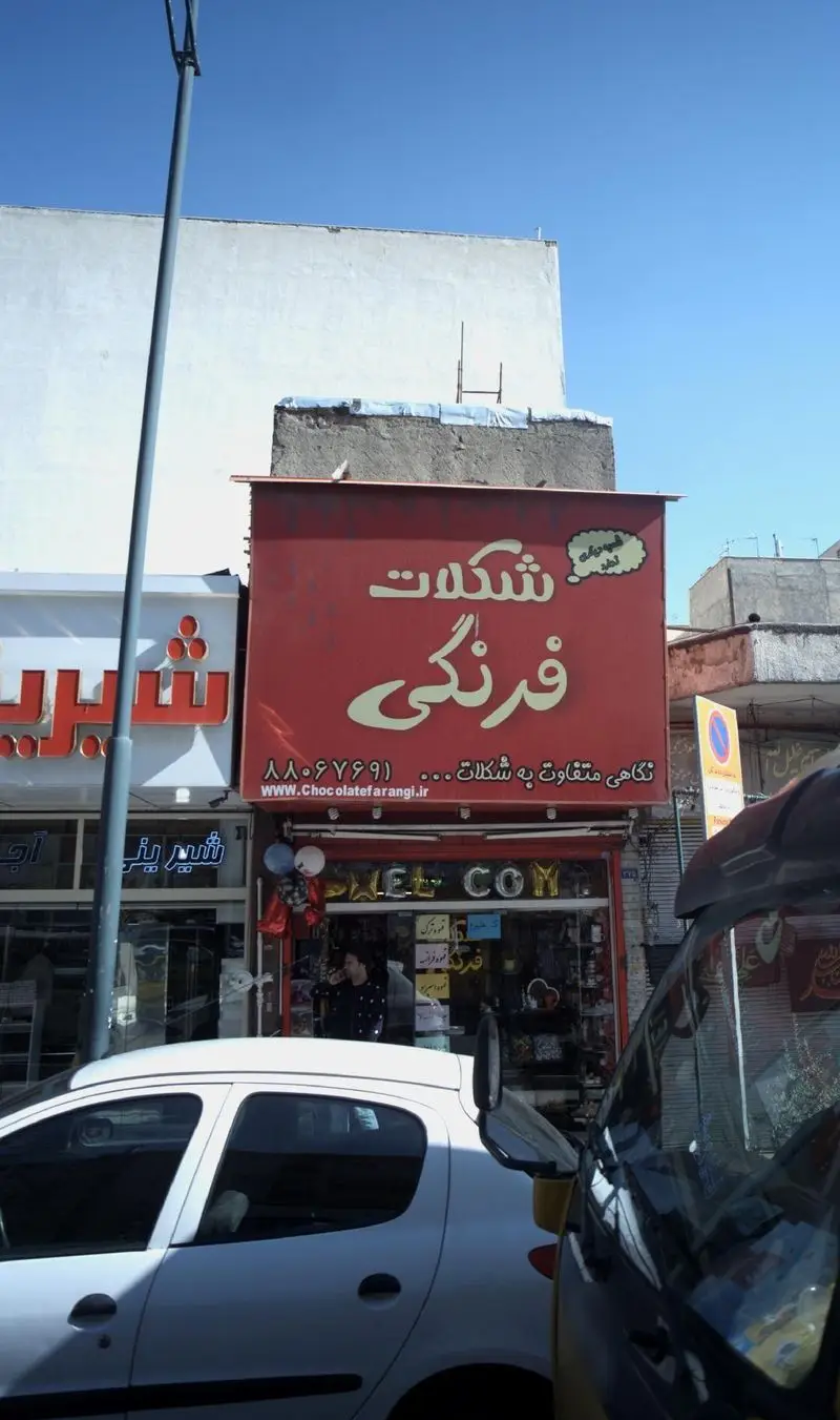 نمای خارجی فروشگاه شکلات فرنگی با تابلوی قرمز بزرگ و شعار "نگاهی متفاوت به شکلات"، آدرس وبسایت Chocolatefarangi.ir و شماره تلفن ۸۸۰۶۷۶۹۱.