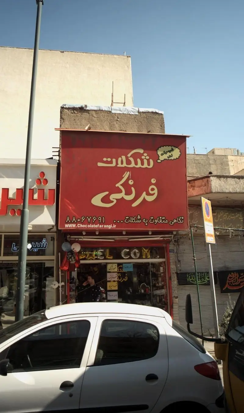 نمای بیرونی فروشگاه شکلات فرنگی با تابلوی قرمز، شماره تماس و وبسایت. ارائه‌دهنده انواع شکلات و شیرینی‌های خاص و متفاوت.