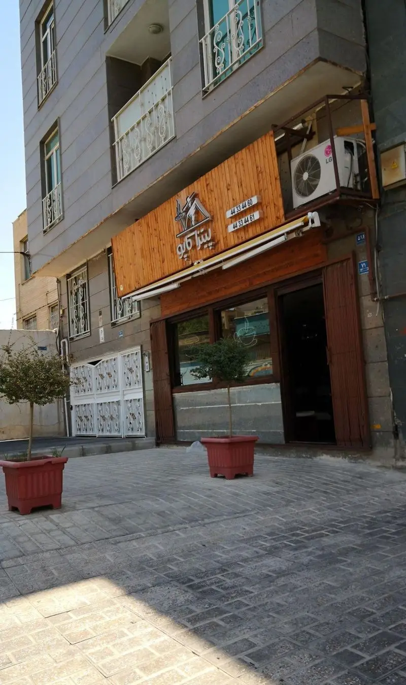 نمای بیرونی و ورودی فست فود کاپو پیتزا با تابلوی چوبی و لوگو، گلدان‌های تزئینی در پیاده‌روی سنگفرش شده جلوی ساختمان.