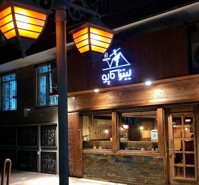 تصویری از فست فود کاپو پیتزا در شب، شامل تابلوی روشن "پیتزا کاپو"، نمای چوبی و آجری، و دو چراغ خیابانی با نور گرم.