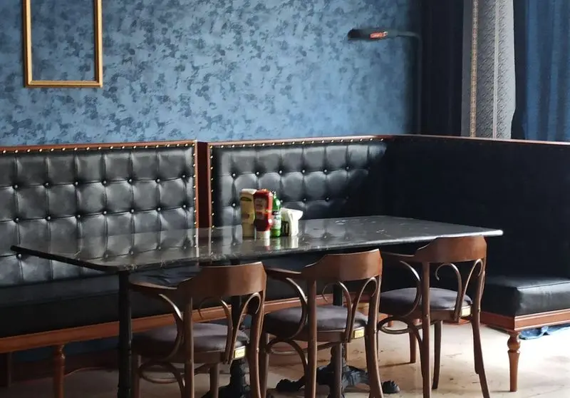 فضای داخلی دنج فست فود گزینگ برگر؛ نیمکت‌های چرمی مشکی، میز بلند مرمر، صندلی‌های چوبی و دیوارهای آبی با سس‌های روی میز.