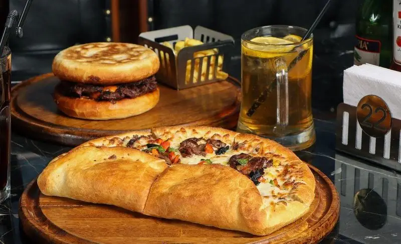 فست فود گزینگ برگر: برگر آبدار، پیتزای گوشت و سبزیجات، سیب زمینی سرخ کرده و نوشیدنی خنک روی میز چوبی.