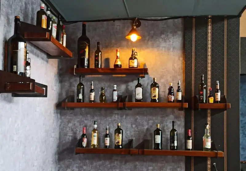 تصویری از قفسه‌های چوبی با بطری‌های متنوع، بخشی از دکوراسیون داخلی جذاب فست فود گزینگ برگر با نورپردازی گرم و دیوار خاکستری.