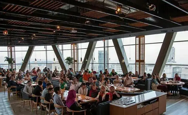 نمای داخلی کافه یا رستوران شیک آوا سنتر، با سقف بلند، پنجره‌های سراسری و افراد زیادی که مشغول دورهمی و معاشرت هستند.