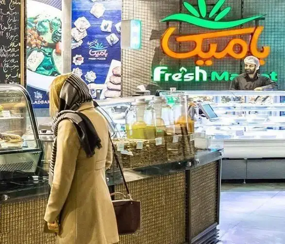 تصویری از مشتری در حال خرید در فروشگاه مدرن Fresh Market بامیکا در آوا سنتر، با ویترین‌های جذاب مواد غذایی تازه، نوشیدنی‌ها و خشکبار.