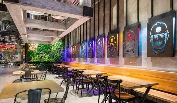 دکوراسیون داخلی رستوران آوا سنتر با میزهای چوبی، دیوارهای سبز طبیعی و تابلوهای نورانی با نقش چهره‌های صنعتی در فضایی آرام و مدرن.