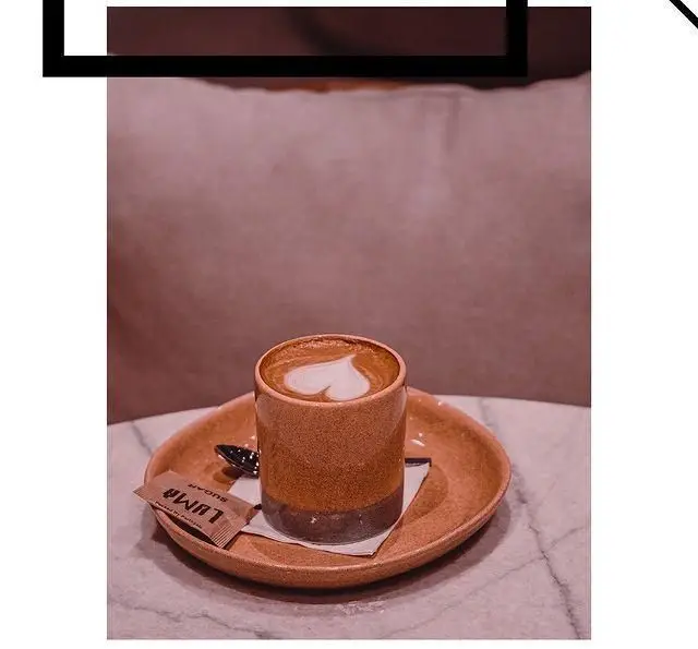 یک فنجان قهوه لاته با لاته آرت طرح قلب، همراه با شکر کافه لوما، سرو شده روی سینی در محیط دلنشین کافه لوما.