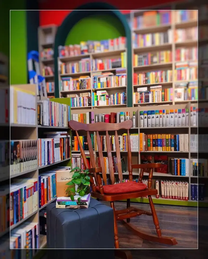 فضای آرام و دنج فروشگاه کتاب داربوک: قفسه‌های بلند پر از کتاب‌های رنگارنگ، صندلی گهواره‌ای چوبی با بالشتک قرمز و پاف با گلدان و کتاب.