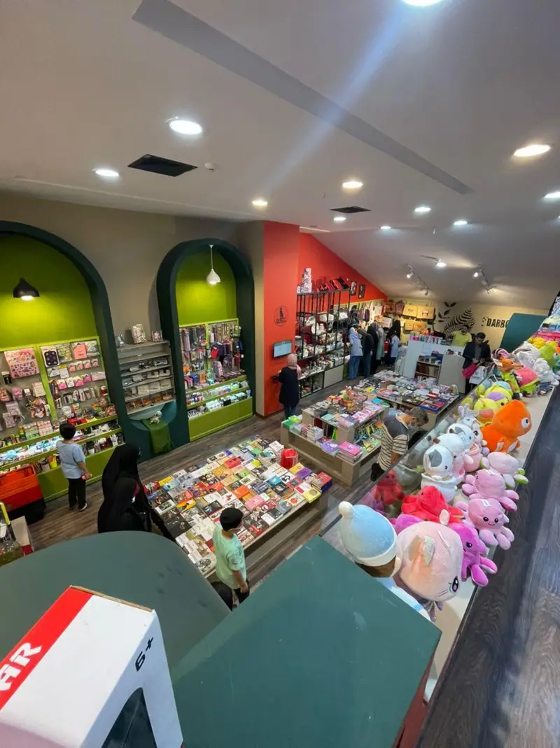 فروشگاه کتاب داربوک: نمای داخلی پر جنب و جوش با مشتریان، قفسه های کتاب رنگارنگ، عروسک ها و لوازم التحریر جذاب برای خرید.