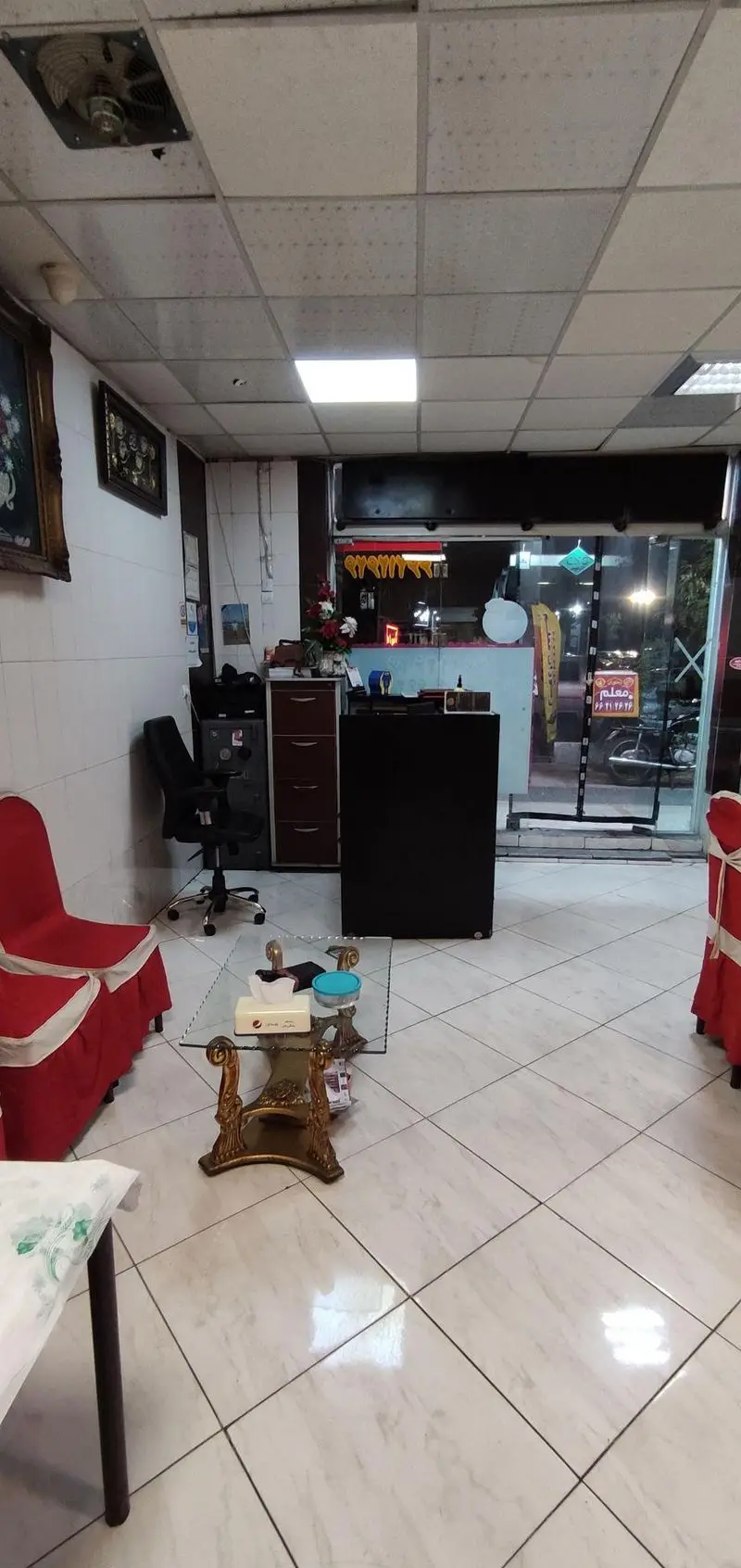 داخلی رستوران معلم با میز و صندلی‌های چیده شده، پیشخوان پذیرش، سقف کاذب و ورودی شیشه‌ای به خیابان، محیطی آرام و تمیز.