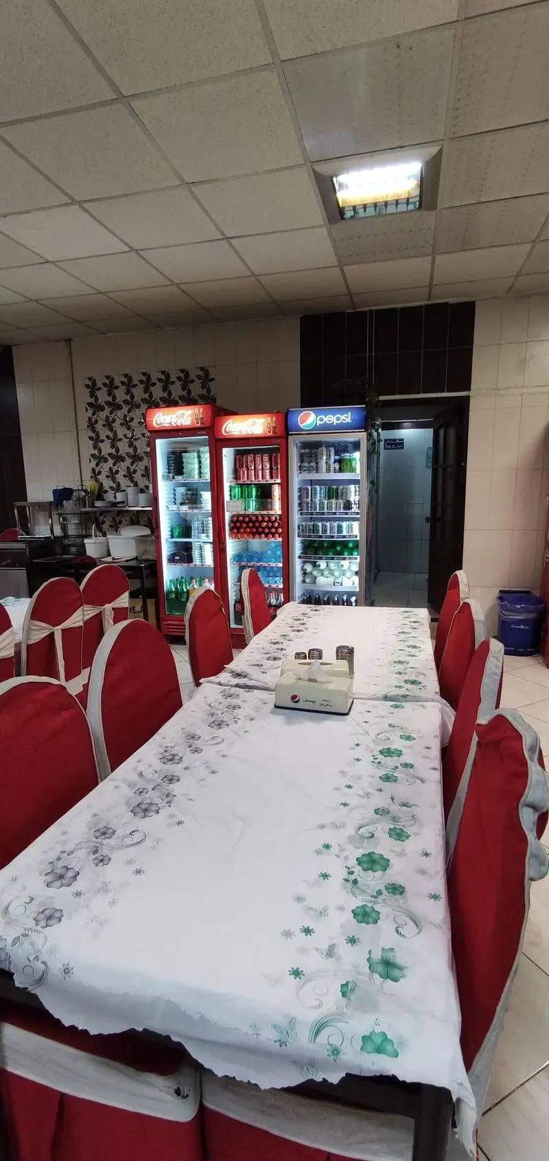 فضای داخلی رستوران معلم با میزهای چیده شده، صندلی‌های قرمز، و یخچال‌های نوشیدنی کوکاکولا و پپسی در محیطی تمیز و آماده پذیرایی.