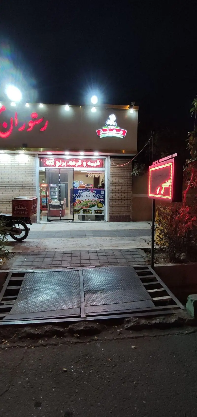 نمای بیرونی رستوران معلم در شب، با تابلوی نئون "قیمه و قورمه" و موتور پیک غذا. شماره تماس‌ها روی شیشه قابل مشاهده است.