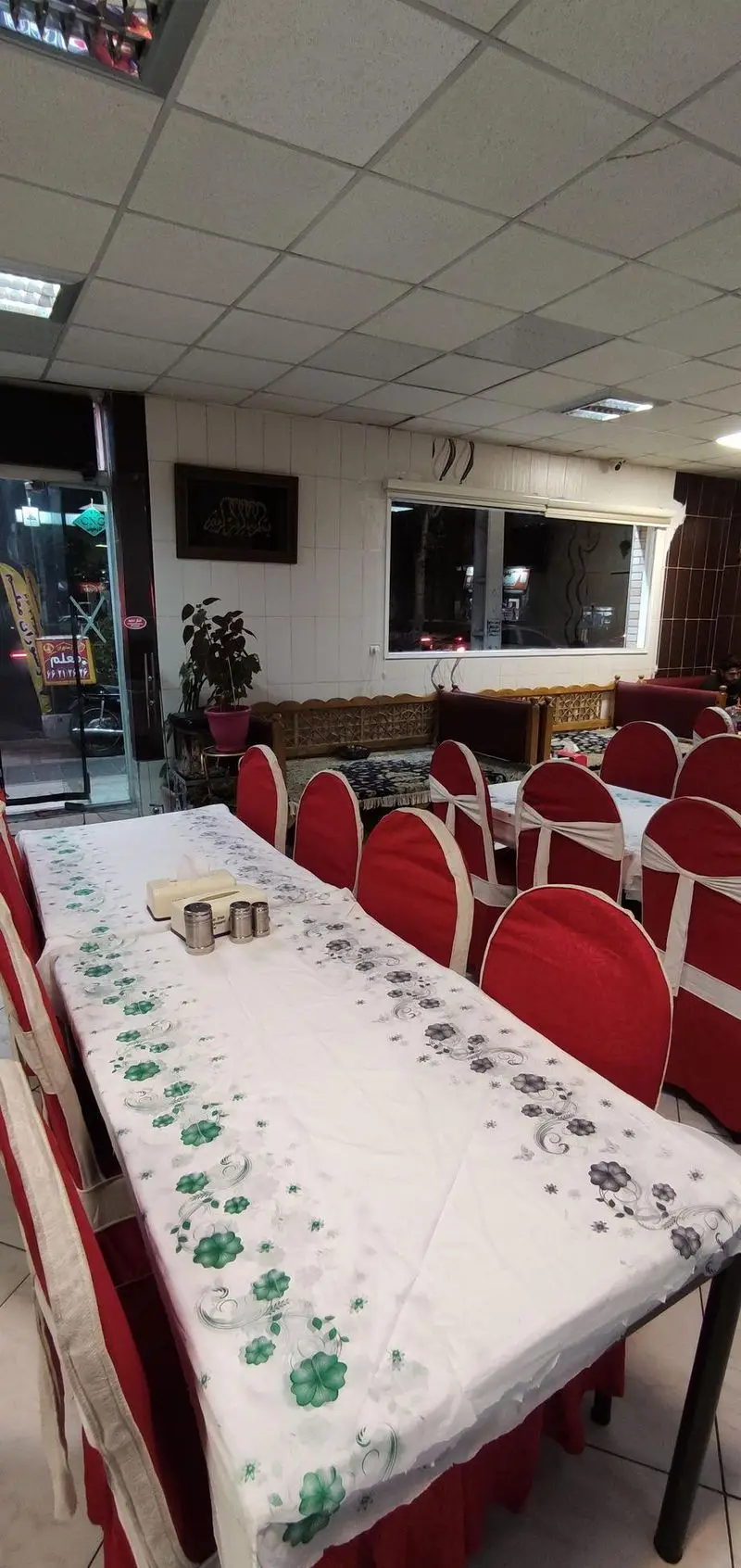 فضای داخلی دلنشین و تمیز رستوران معلم در شب، شامل میزهای آماده با رومیزی طرح‌دار، صندلی‌های قرمز و بخش سنتی جهت پذیرایی.