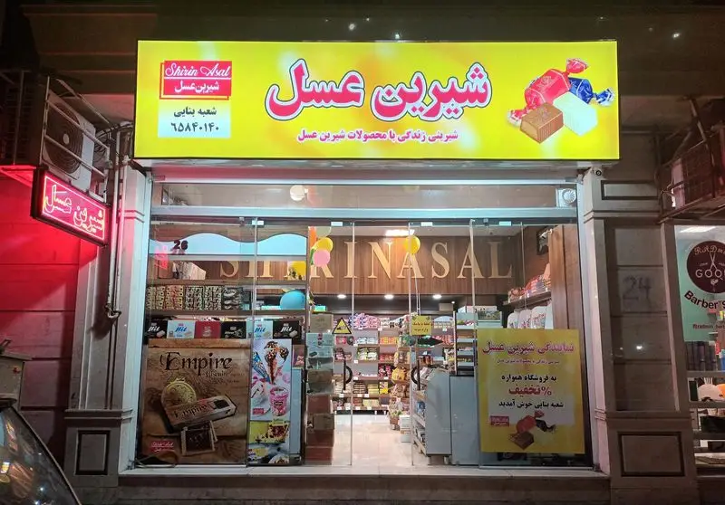 نمای شب فروشگاه شیرین عسل بنایی با تابلوی روشن و ویترین پر از انواع شیرینی، شکلات و محصولات لذیذ شیرین عسل برای مشتریان.