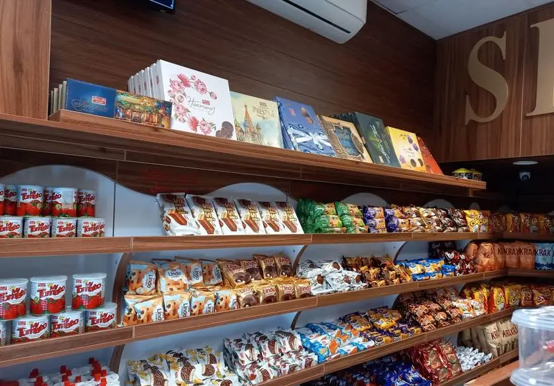 تصویر داخلی فروشگاه شیرین عسل بنایی با قفسه‌های چوبی پر از محصولات، تافی، ویفر، شکلات، بیسکوییت و جعبه‌های هدیه.