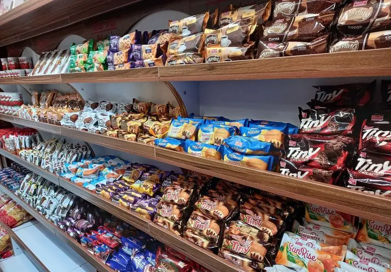 قفسه‌های چوبی فروشگاه شیرین عسل بنایی پر از انواع کیک، کلوچه، بیسکویت و شیرینی‌های بسته‌بندی از برندهای معروف مانند میلانو، مورنینگ، تینای و سانرایز.