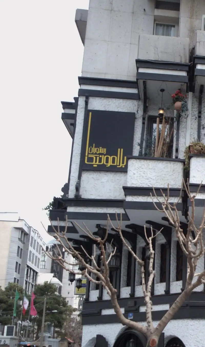 نمای بیرونی رستوران ایتالیایی بلامونیکا با تابلوی طلایی رنگ، معماری سیاه و سفید زیبا و بالکن‌های تزئین شده، در یک روز ابری.