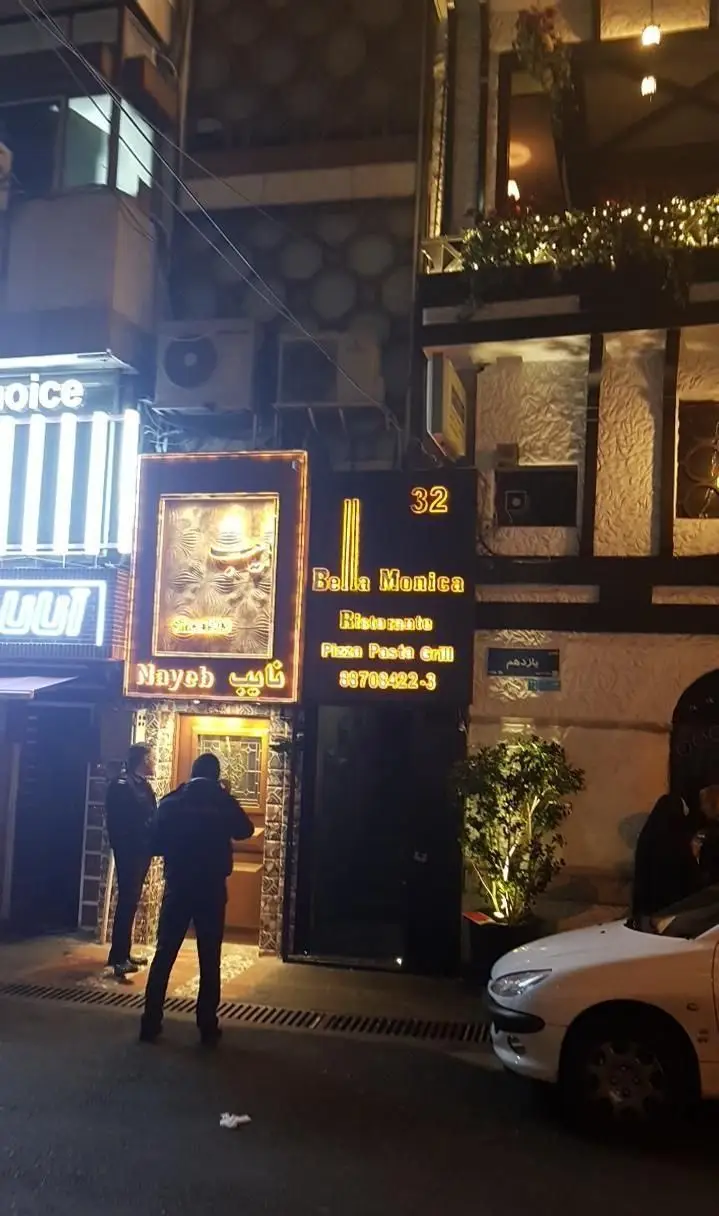 تصویر شبانه از نمای بیرونی مدرن و شیک رستوران ایتالیایی بلامونیکا، همراه با تابلوهای نوری پیتزا، پاستا و گریل برای مشتریان.