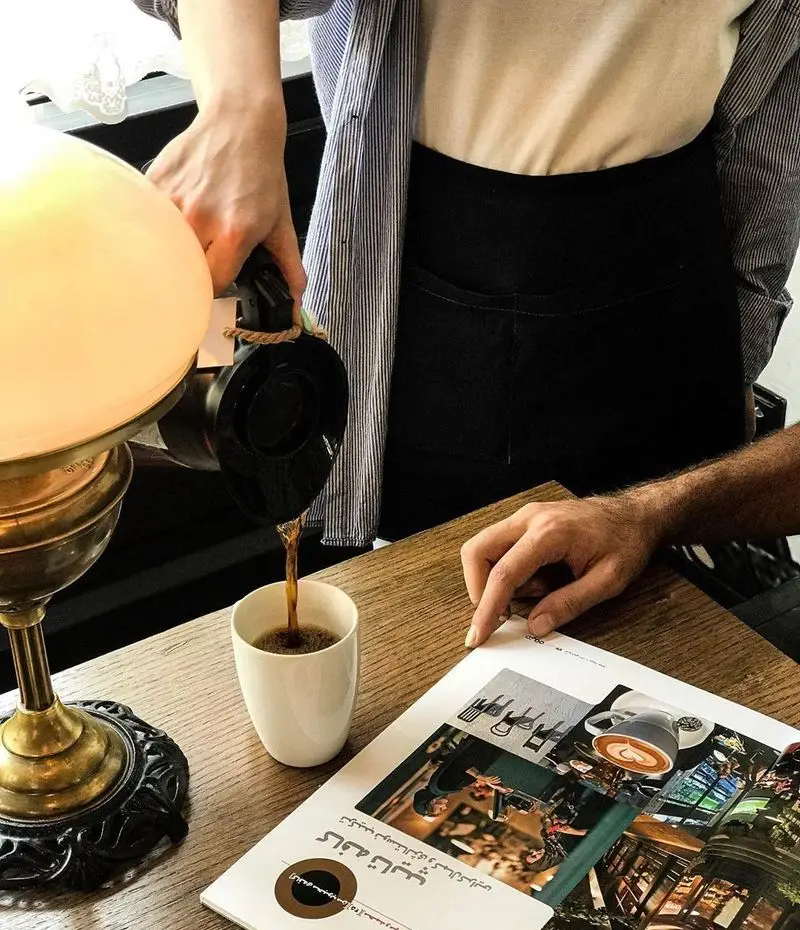 لحظه‌ای دلنشین در کافه تایپ: باریستا قهوه را با دقت در ماگ سفید می‌ریزد، کنار مجله‌ای از کافه و چراغ رومیزی بر میز چوبی.