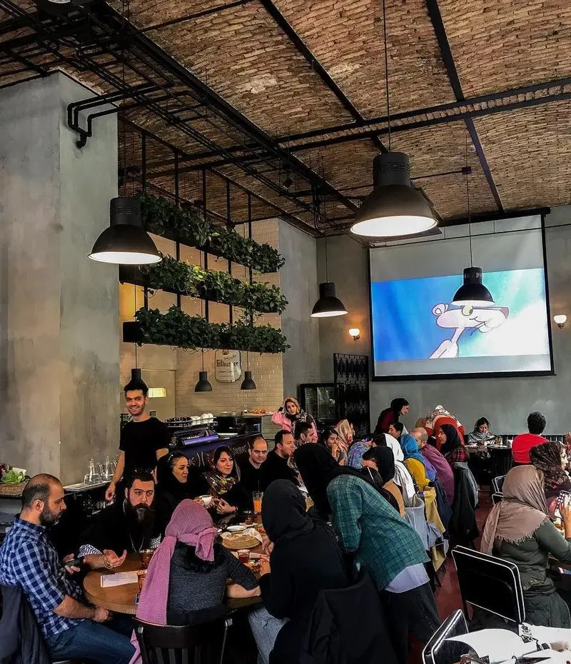 تصویری از کافه تایپ، فضایی شلوغ و دوستانه با سقف آجری، نورپردازی صنعتی و دیوار سبز. مشتریان از دورهمی و نمایش کارتون لذت می‌برند.