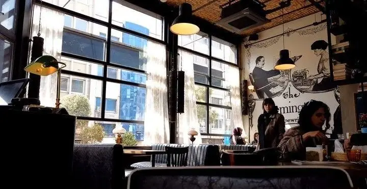 محیط دنج کافه تایپ با نورپردازی گرم، مبلمان مدرن، پنجره‌های بزرگ و دیوارکوبی جذاب از ریمینگتون تایپ‌رایتر و فضای مناسب مطالعه و نوشتن.