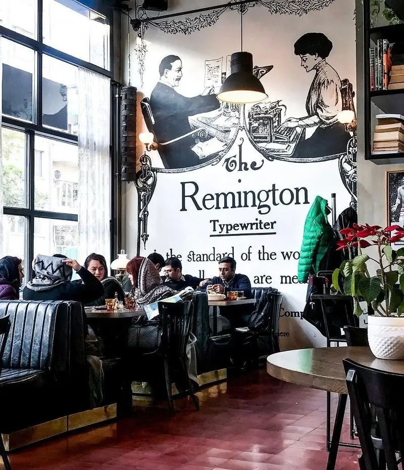 نمایی دنج از کافه تایپ با دیوارنگاره بزرگ ماشین تحریر ریمینگتون، مشتریان در حال نوشیدن و گفتگو در فضای کلاسیک و دلنشین.