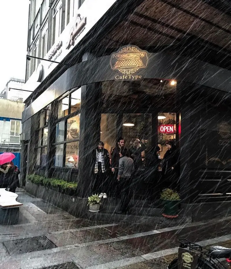 نمایی جذاب از کافه تایپ در هوای بارانی، با سردر مشکی و لوگوی کلاسیک ماشین تحریر. تابلوی 'OPEN' به رنگ قرمز و مشتریان در ورودی منتظر.