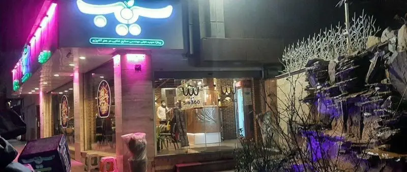 تصویر جذاب شب از نمای بیرونی شعبه پیتزا سیب ۳۶۰، رستورانی مدرن با تابلوی نئون درخشان، ورودی شیشه‌ای و آبنمای تزئینی با نور بنفش.