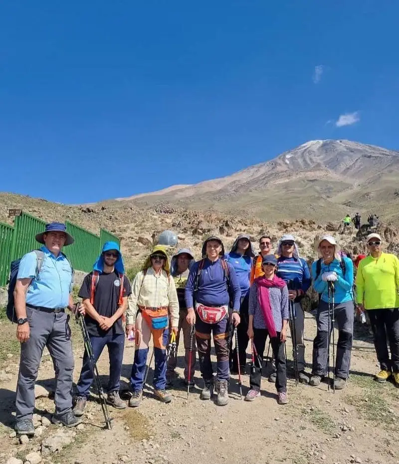 گروه کوهنوردان شاد در دامنه کوهی باشکوه و برفی زیر آسمان آبی، نمادی از ماجراجویی و آرامش. کافه فرش، مکانی ایده‌آل برای استراحت پس از تجربه‌ای مشابه.