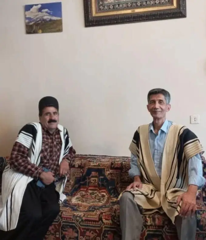 کافه فرش: دو مرد با لباس بومی بر روی کاناپه‌ای با طرح گلیم و قالی سنتی ایرانی نشسته‌اند که نمادی از فرهنگ و اصالت است.