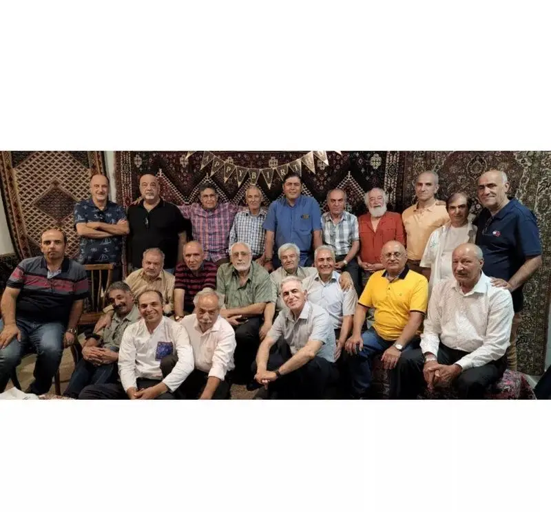 تصویری از جمع دوستانه مردان مسن در کافه فرش، با پس‌زمینه‌ای از فرش‌های دستباف نفیس ایرانی، در حال برگزاری جشن تولد.