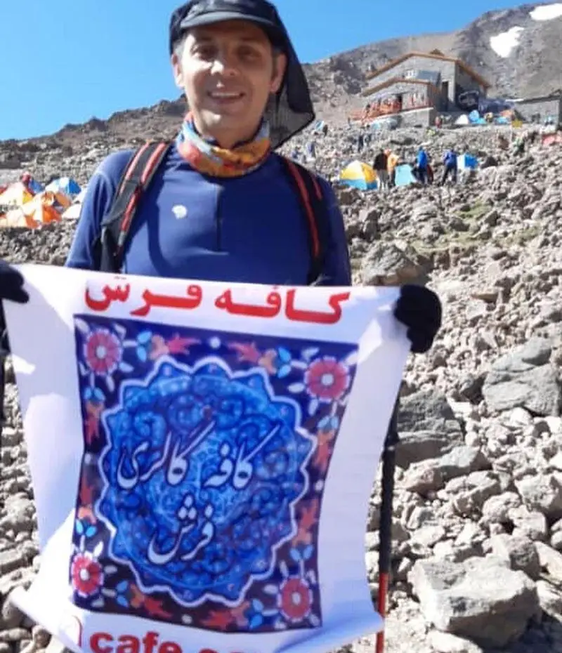 تصویر مردی در لباس کوهنوردی با کلاه و کوله پشتی که بنر بزرگ کافه فرش و لوگوی آبی آن را در دست دارد، در محیط کوهستانی با پناهگاه و چادرهای کوهنوردی.