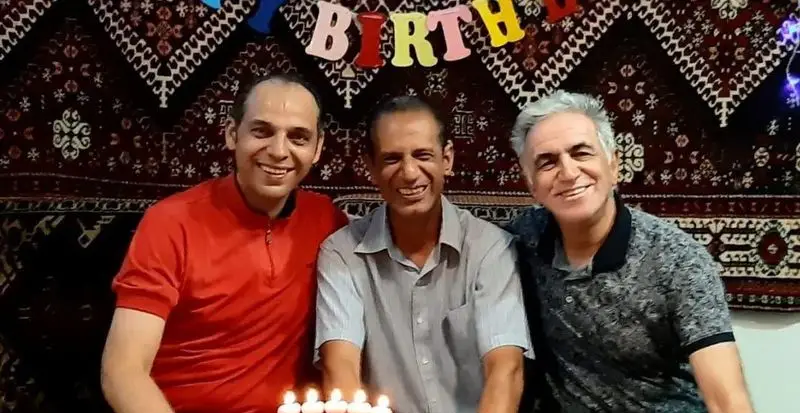 سه مرد شاد در حال جشن تولد در فضای دلنشین و گرم کافه فرش با پس‌زمینه فرش اصیل ایرانی و طرح‌های سنتی زیبا.