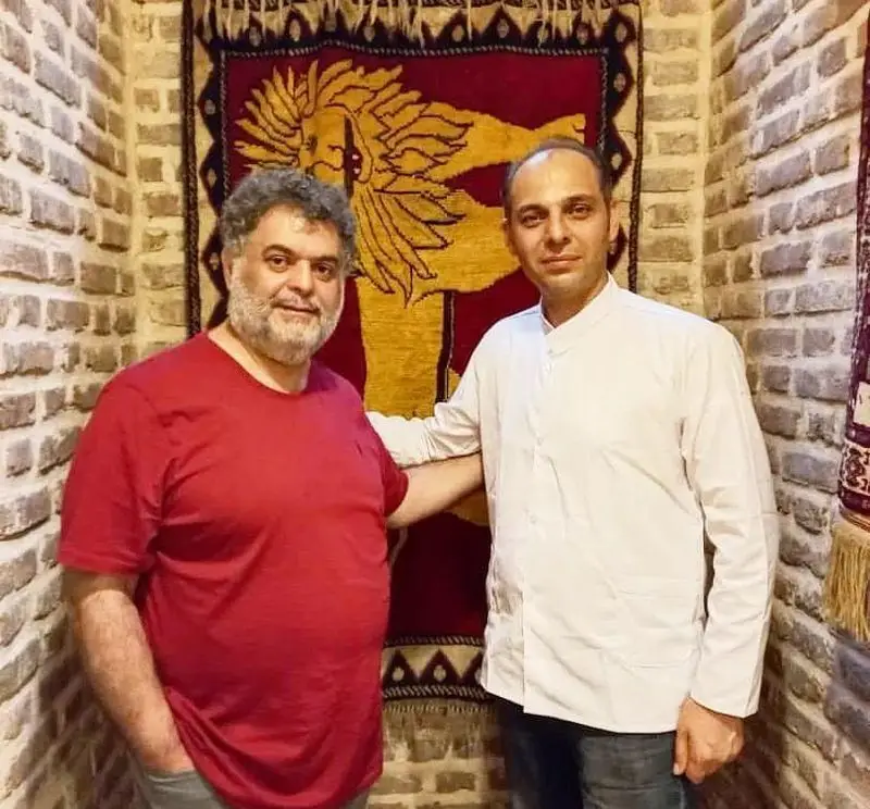 دو مرد در کافه فرش، لذت بردن از فضای دلنشین و دنج با پس‌زمینه دیوار آجری و تابلوفرش اصیل ایرانی با طرح شیر.